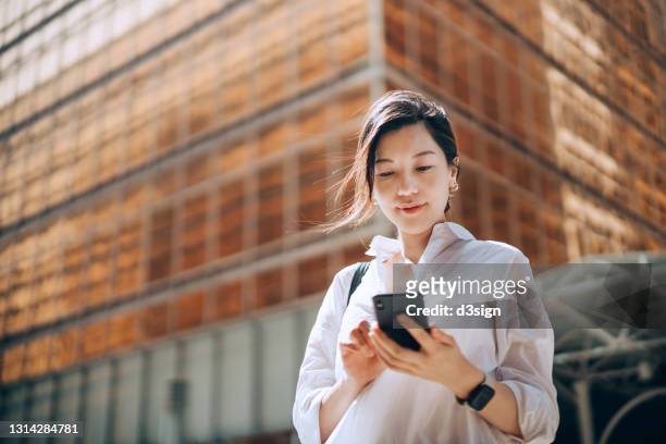 low angle portrait of young asian businesswoman checking emails on smartphone outside office building in financial district. with contemporary corporate skyscrapers in background. making business connections throughout the city - ringing stockfoto's en -beelden
