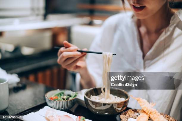 close up mid-section of young asian woman enjoying a bowl of japanese style udon soup noodles, with assorted sushi and seafood tempura freshly served on the dining table in a restaurant. asian cuisine and food. eating out lifestyle - udon noodle stock pictures, royalty-free photos & images