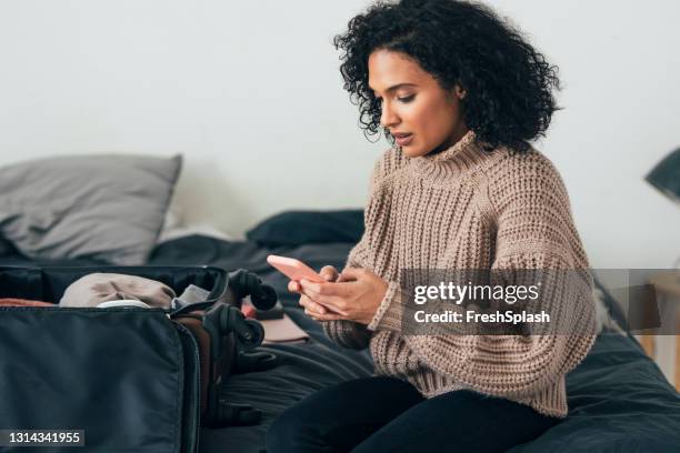 beautiful young woman packing her suitcase for winter holiday and booking a flight online on her mobile phone - female worried mobile imagens e fotografias de stock