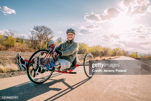 joven guapo sonriendo y mirando a la cámara mientras está sentado en su ciclo de mano rojo usando aparatos ortopédicos para el ejercicio adaptativo al aire libre al atardecer con espacio de copia - assistive technology fotografías e imágenes de stock