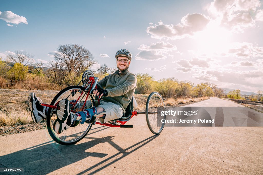 Knappe Jonge Mens die en de Camera glimlacht bekijkt terwijl het Zitten op Zijn Rode Handcycle die Orthetische Beensteunen draagt voor Adaptieve Oefening in openlucht bij Zonsondergang met Exemplaarruimte