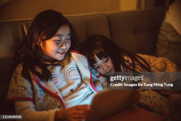 lovely little sisters using digital tablet together joyfully on the sofa at home at night - kid leaning stock pictures, royalty-free photos & images