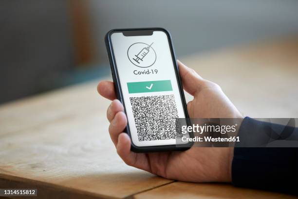 man holding mobile phone with digital vaccination card over wooden table - smartphone stock-fotos und bilder