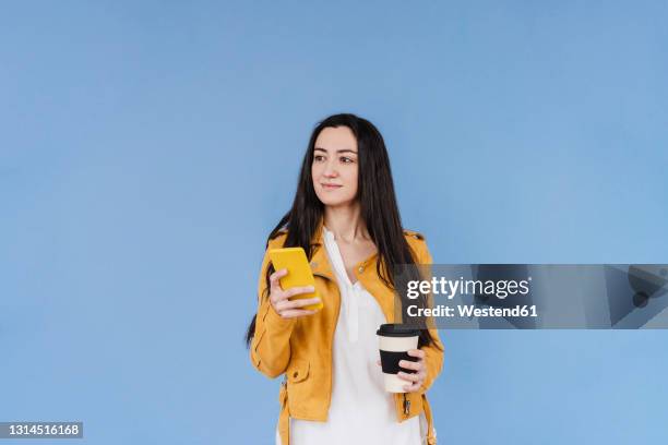 smiling woman with mobile phone looking away while standing against blue background - woman smiling white background stock-fotos und bilder