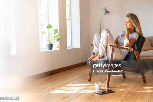 smiling woman with hand on chin looking away while sitting on chair at home - comfortable fotografías e imágenes de stock