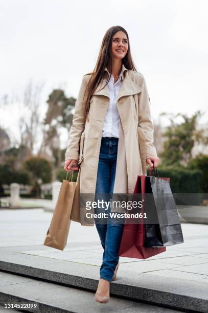 woman in overcoat moving down from stairs while holding shopping bags - überzieher stock-fotos und bilder