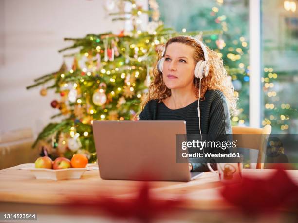 woman with headphones looking away while sitting in front of laptop at christmas - christmas music listen stock pictures, royalty-free photos & images