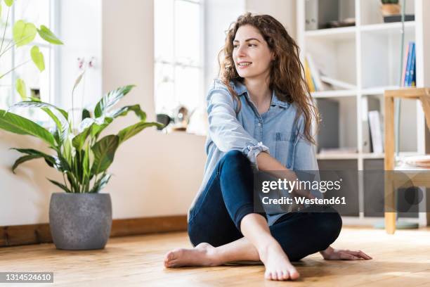 smiling woman with long hair looking away while sitting on floor in living room - sitting on floor stock pictures, royalty-free photos & images