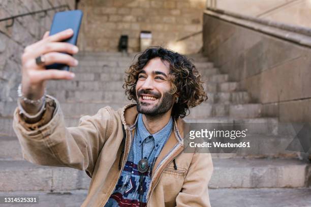 happy hipster man taking selfie through mobile phone while sitting on steps - dyed red hair fotografías e imágenes de stock