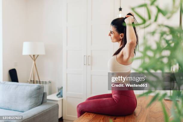 mature woman tying ponytail while sitting on table in living room - soutien fotografías e imágenes de stock