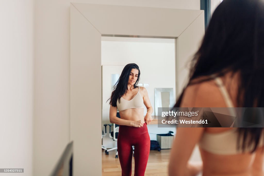 Woman looking in mirror while adjusting pants in living room at home