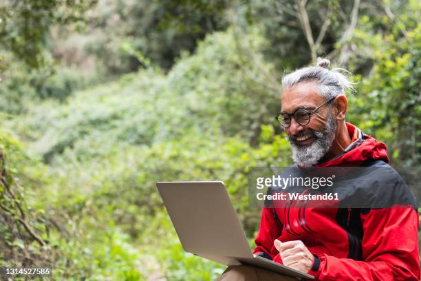 smiling senior man using laptop while sitting in forest - man bun stock pictures, royalty-free photos & images