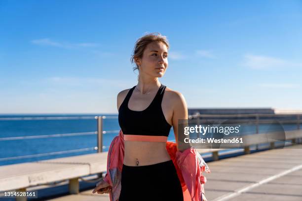 beautiful female jogger removing jacket while looking away during sunny day - uitkleden stockfoto's en -beelden