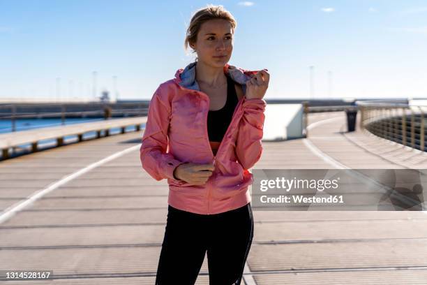 female athlete unzipping jacket while standing on boardwalk - entkleiden stock-fotos und bilder