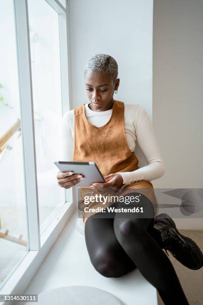 serious woman looking at digital tablet while sitting by window in living room - women wearing black stockings stock pictures, royalty-free photos & images