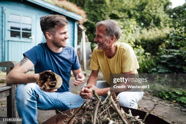 son holding firewood discussing with father while crouching in backyard - wooden shed stock pictures, royalty-free photos & images