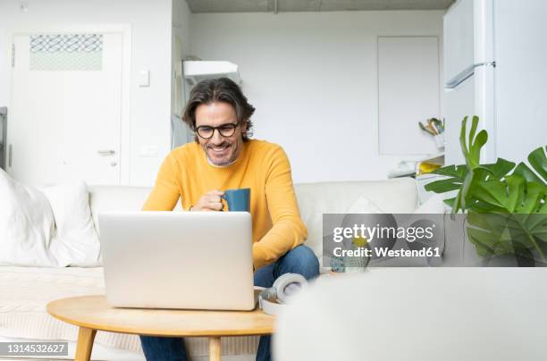 smiling man holding mug while using laptop at home - man middelbare leeftijd woonkamer stockfoto's en -beelden