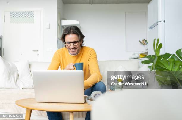 smiling man holding mug while using laptop at home - homem computador imagens e fotografias de stock