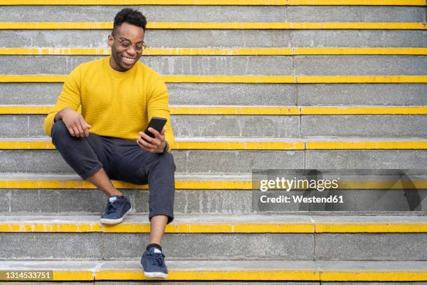 smiling young man using smart phone while sitting on staircase - man stairs sitting stock-fotos und bilder