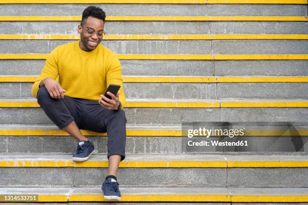smiling young man using smart phone while sitting on staircase - man sitting alone stock pictures, royalty-free photos & images