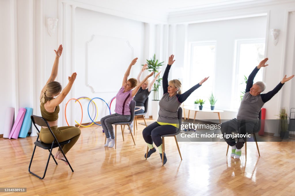 Yoga instructor showing new relaxation exercise to senior people during yoga class with chairs