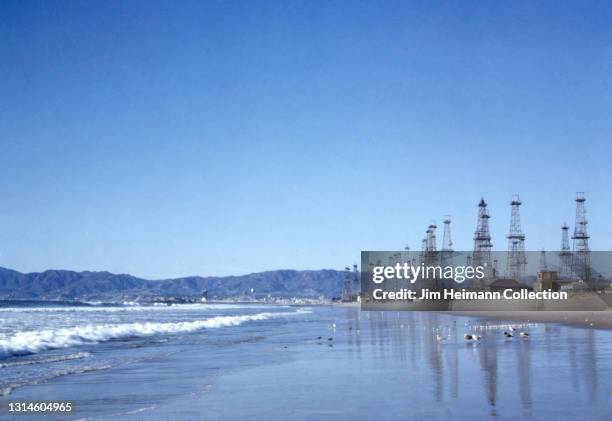 35mm film photo shows the beach at Venice, California during high tide. Birds walk across the sand, and oil derricks are lined up along the coast as...