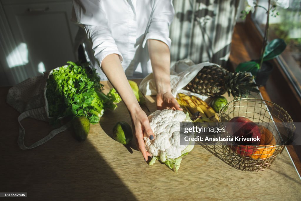 Beautiful woman unpacks a full fabric bag with fruits and vegetables on the kitchen.