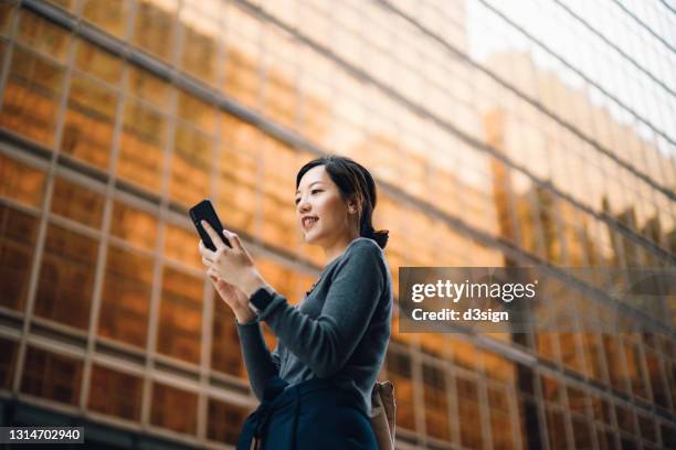 low angle portrait of young asian businesswoman managing online banking with mobile app on smartphone on the go. transferring money, paying bills, checking balance in the city against corporate skyscrapers. making business connections throughout the city - aktienkauf stock-fotos und bilder