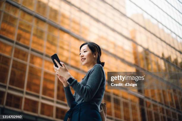 low angle portrait of young asian businesswoman managing online banking with mobile app on smartphone on the go. transferring money, paying bills, checking balance in the city against corporate skyscrapers. making business connections throughout the city - 電話　ビジネス ストックフォトと画像