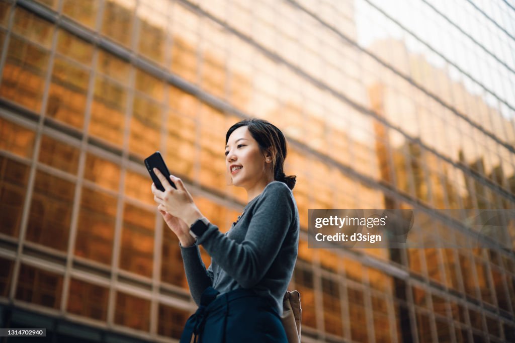 Low angle portrait of young Asian businesswoman managing online banking with mobile app on smartphone on the go. Transferring money, paying bills, checking balance in the city against corporate skyscrapers. Making business connections throughout the city