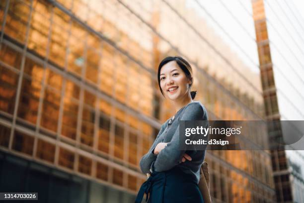 low angle portrait of confident and successful young asian businesswoman standing against contemporary corporate skyscrapers in financial district. female leadership and determined to success - central asia stock-fotos und bilder