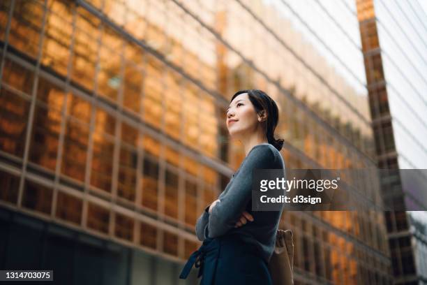 low angle portrait of confident and successful young asian businesswoman standing against contemporary corporate skyscrapers in financial district. female leadership and determined to success - skyscraper film stock-fotos und bilder