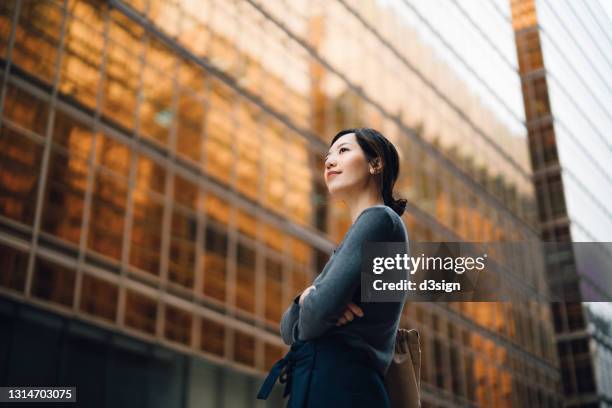 low angle portrait of confident and successful young asian businesswoman standing against contemporary corporate skyscrapers in financial district. female leadership and determined to success - skyscraper foto e immagini stock