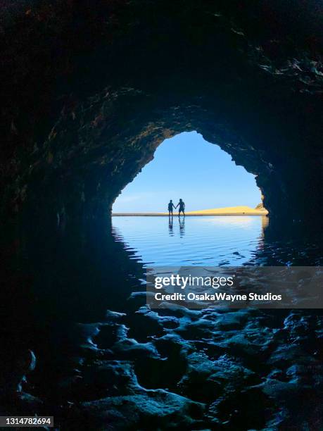 pair of people holding hands and standing on beach in front of sea coast cave. - kapaa beach park stock pictures, royalty-free photos & images