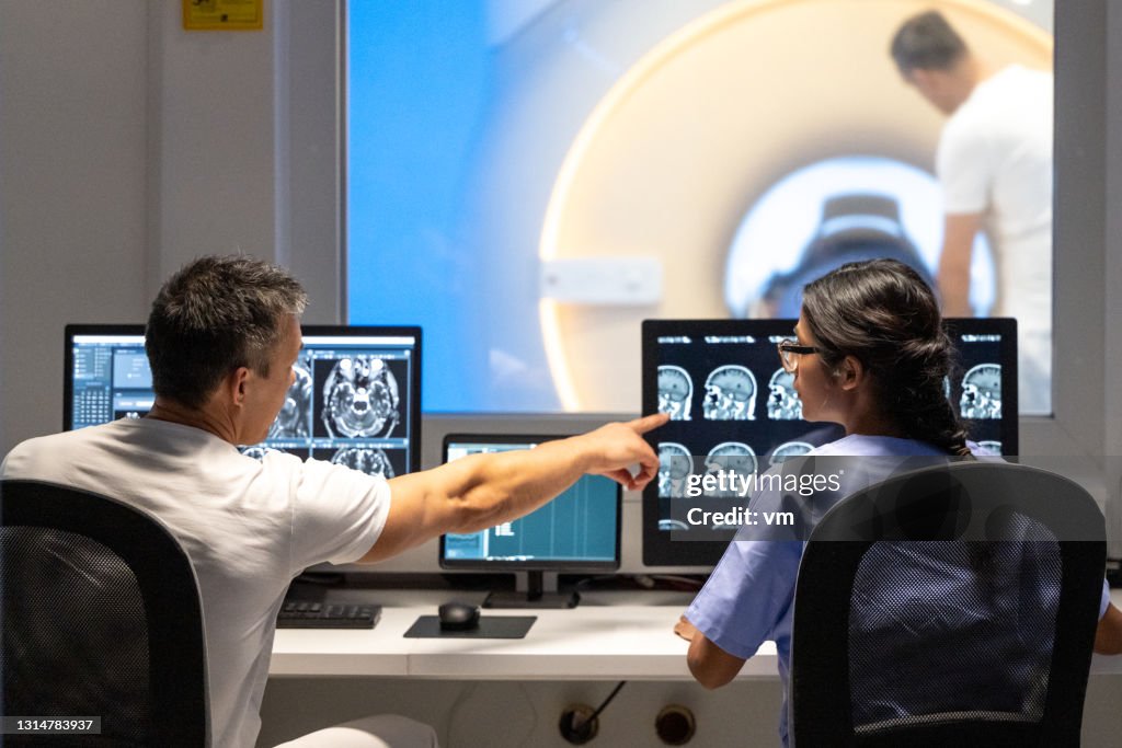 Back view of two doctors analyzing MRI scan results. Man pointing at images on monitor screen.