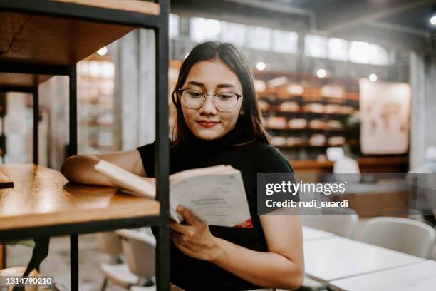 jonge vrouwelijke student die boek van plank in bibliotheek bij communautaire universiteit kiest - bibliotheek stockfoto's en -beelden