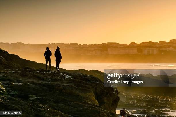 People standing on rocks silhouetted by the setting sun at Little Fistral in Newquay in Cornwall.