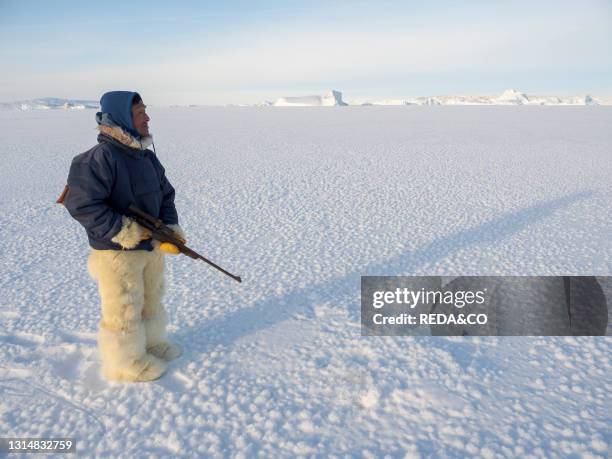 Watching a breathing hole of a seal. Inuit hunter wearing traditional trousers and boots made from polar bear fur on the sea ice of the Melville Bay...