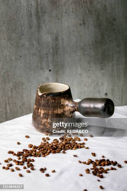 Brown craft ceramic coffee pot Turkish cezve standing on white tablecloth with coffee beans above. Gray wall behind. Copy space.