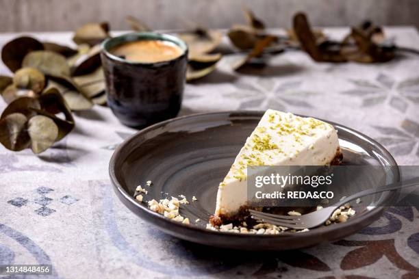 Piece of raw vegan cheesecake. No bake gluten free. Decorated by lime zest and cashew nuts on plate. Cup of black coffee and Eucalyptus branches on...