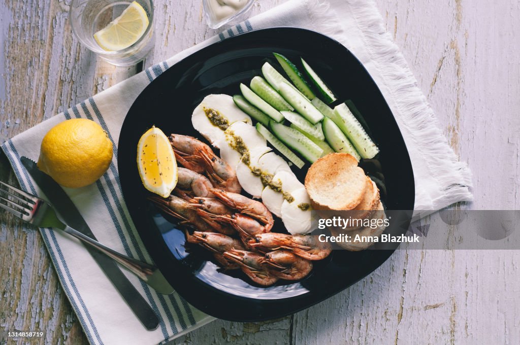 Plate of delicious appetizer: fried shrimp, mozzarella cheese, pesto, fried bread, lemon and cucumber sticks with sauce on gray wooden table.