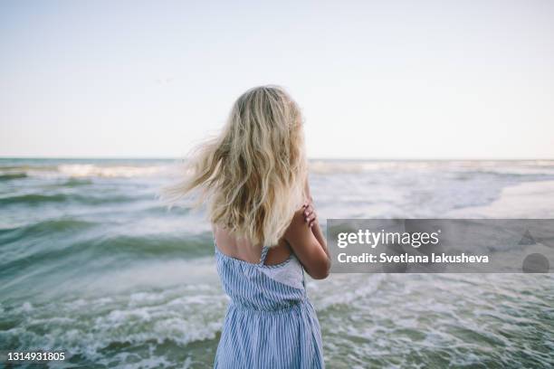 a young blonde woman in a blue sundress walking along the seashore and enjoys the wind and the sea - mujeres de mediana edad fotografías e imágenes de stock
