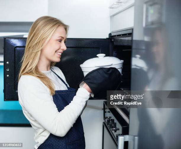 schöne junge blonde frau an offener ofentür hält eine auflaufschale mit ofenhandschuhen in der küche - auflaufgericht stock-fotos und bilder
