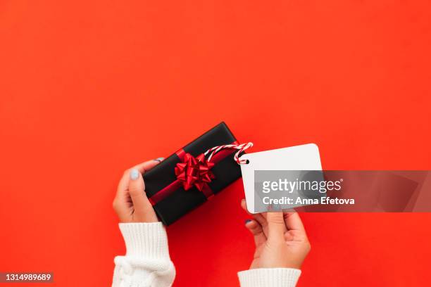 black gift box with red bow and white tag in woman hands on red background. black friday shopping concept. copy space for your design - gift box tag stockfoto's en -beelden