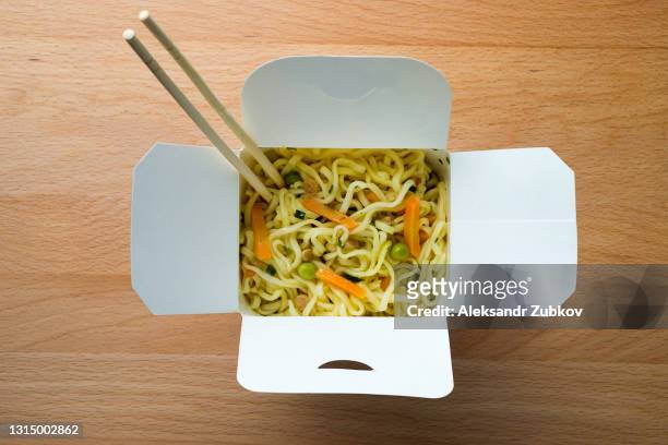 cooked instant noodles, sprinkled with spices, vegetables and herbs, freeze-dried meat. nearby are wooden chopsticks. asian chicken soup with ramen noodles in a paper box, on the dining room kitchen table. the concept of home delivery of food. - chinese noodles stockfoto's en -beelden