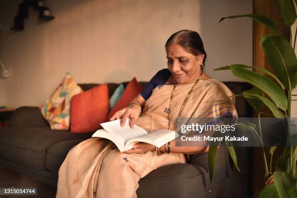 senior woman reading a book sitting on a sofa - indian old woman stock-fotos und bilder