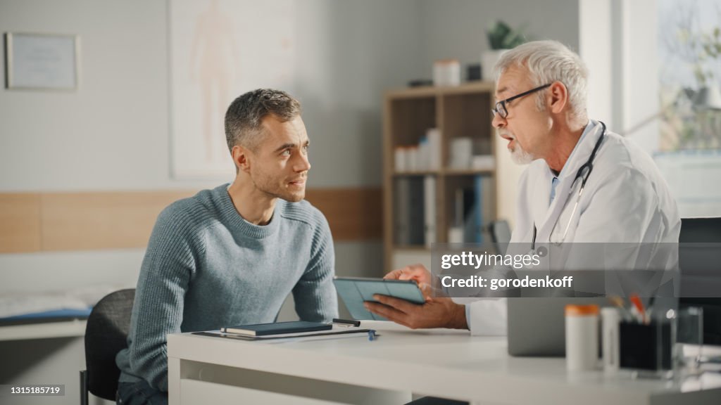 Experienced Middle Aged Family Doctor Showing Analysis Results on Tablet Computer to Male Patient During Consultation in a Health Clinic. Physician Sitting Behind a Desk in Hospital Office.