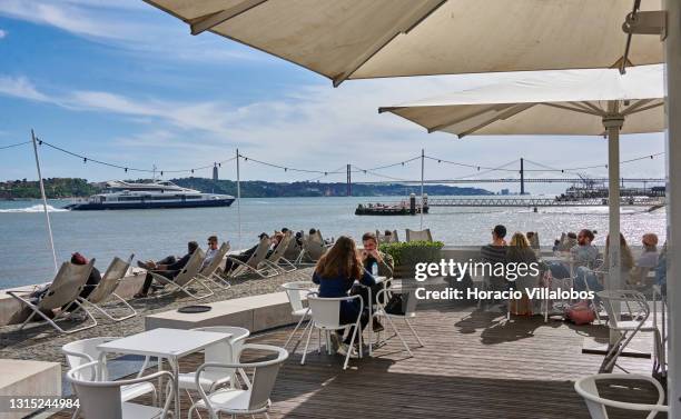People sit at outdoor tables and lounge chairs in Quiosque Ribeira das Naus by the Tagus River a day before the end of the state of emergency during...