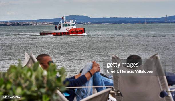Small tugboat sails the Tagus River as patrons sit at lounge chairs in Quiosque Ribeira das Naus by the Tagus River a day before the end of the state...