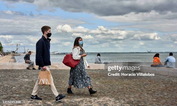 Amask-clad couple walks near Quiosque Ribeira das Naus by the Tagus River carrying a Zara shopping bag a day before the end of the state of emergency...